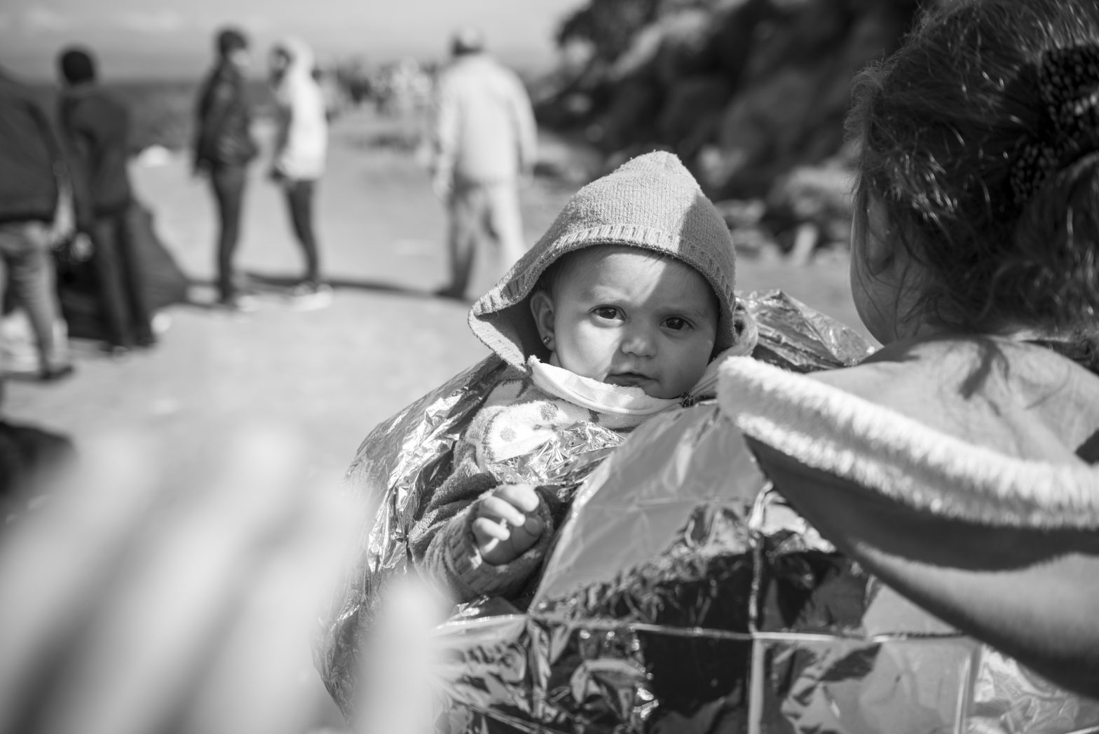 Lesbos, Greece - October 25, 2015: Ala, 10 months old and from Qamishli, Syria, is wrapped in a foil blanket after reaching the coast of Lesbos at the end of a dangerous boat ride from Turkey. The family is Kurdish. Other refugees are in the background. The hand of another refugee is in the foreground, waving at Ala.