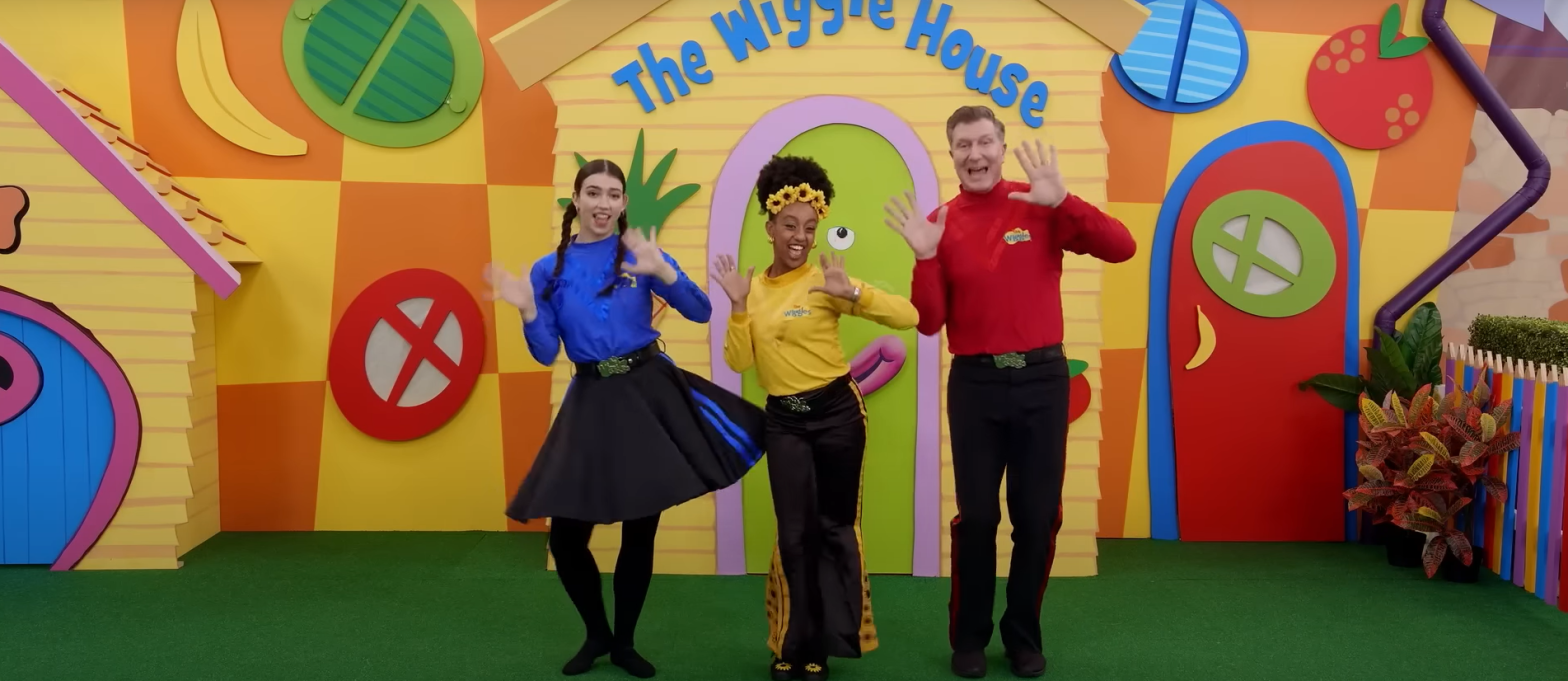 Blue, yellow and red members of the Wiggles dancing in front of The Wiggle House